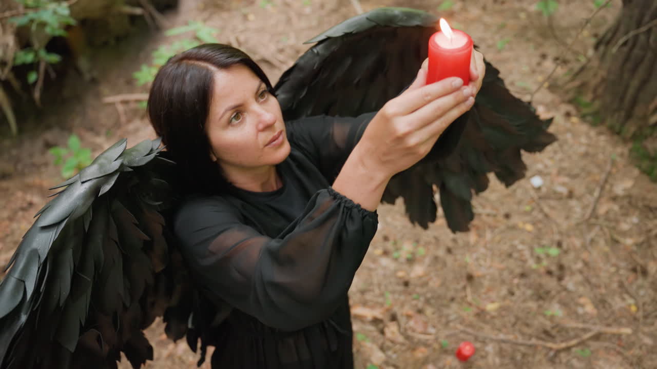 Top view of woman dressed in black with large black wings holding burning red candle raised in offering during mystical forest ritual, symbolizing connection with spirits