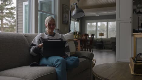 Woman-sitting-on-the-couch-in-front-of-a-bay-window,-drinking-coffee-while-scrolling-on-a-tablet-with-her-feet-on-the-ground