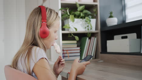 Un-Adolescente-Con-Auriculares-Usa-Un-Teléfono-Inteligente,-Come-Helado-En-Su-Habitación