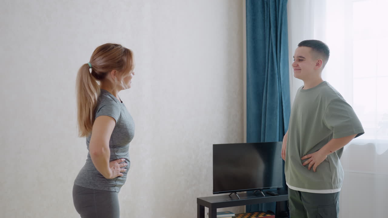 Smiling female gym instructor guides teen boy through stretching posture in bright room with TV and curtain in background, both holding hips and engaging in light exercise