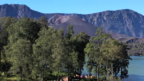 Campsite-with-colorful-tents-at-shores-of-crater-lake-of-Mount-Rinjani-Volcano-Indonesia,-Nusa-Tenggara,-Aerial-pedestal-rising-shot