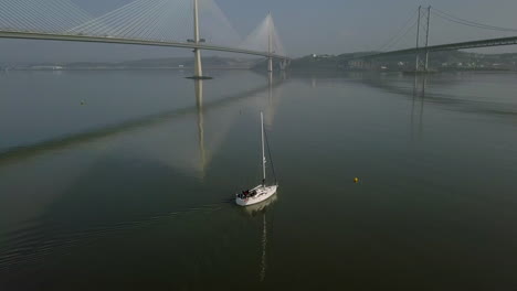 aerial footage of a sailing boat travelling between the forth bridges at south queensferry on a sunny day in west lothian, scotland