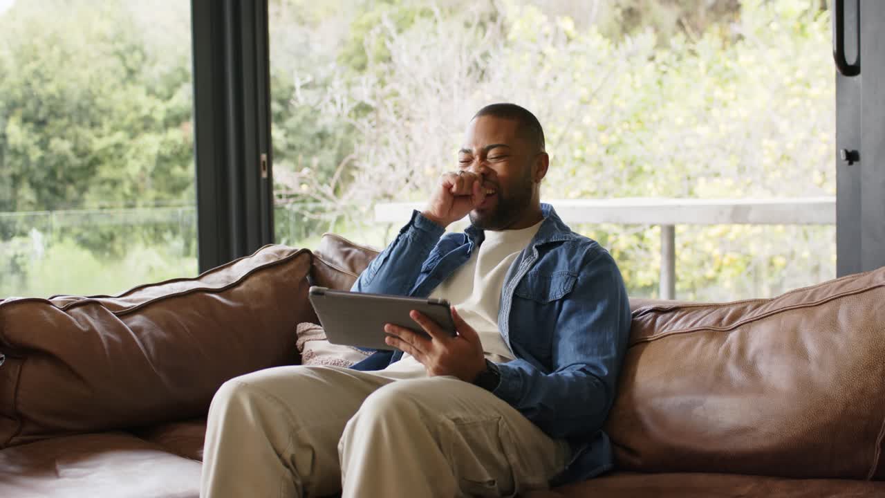 African American man pausing on sofa by glass door tapping tablet scrolling, coughing mid-scroll