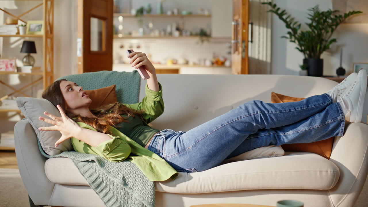 Donna positiva che effettua una videochiamata con lo smartphone, distesa sul divano a casa. Signora che parla