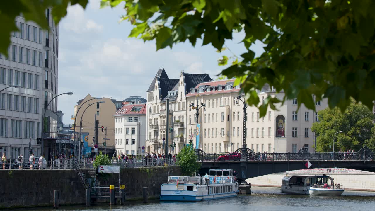 Tour boats glide along Spree River, framed by leafy branches, historic Berlin architecture, daylight