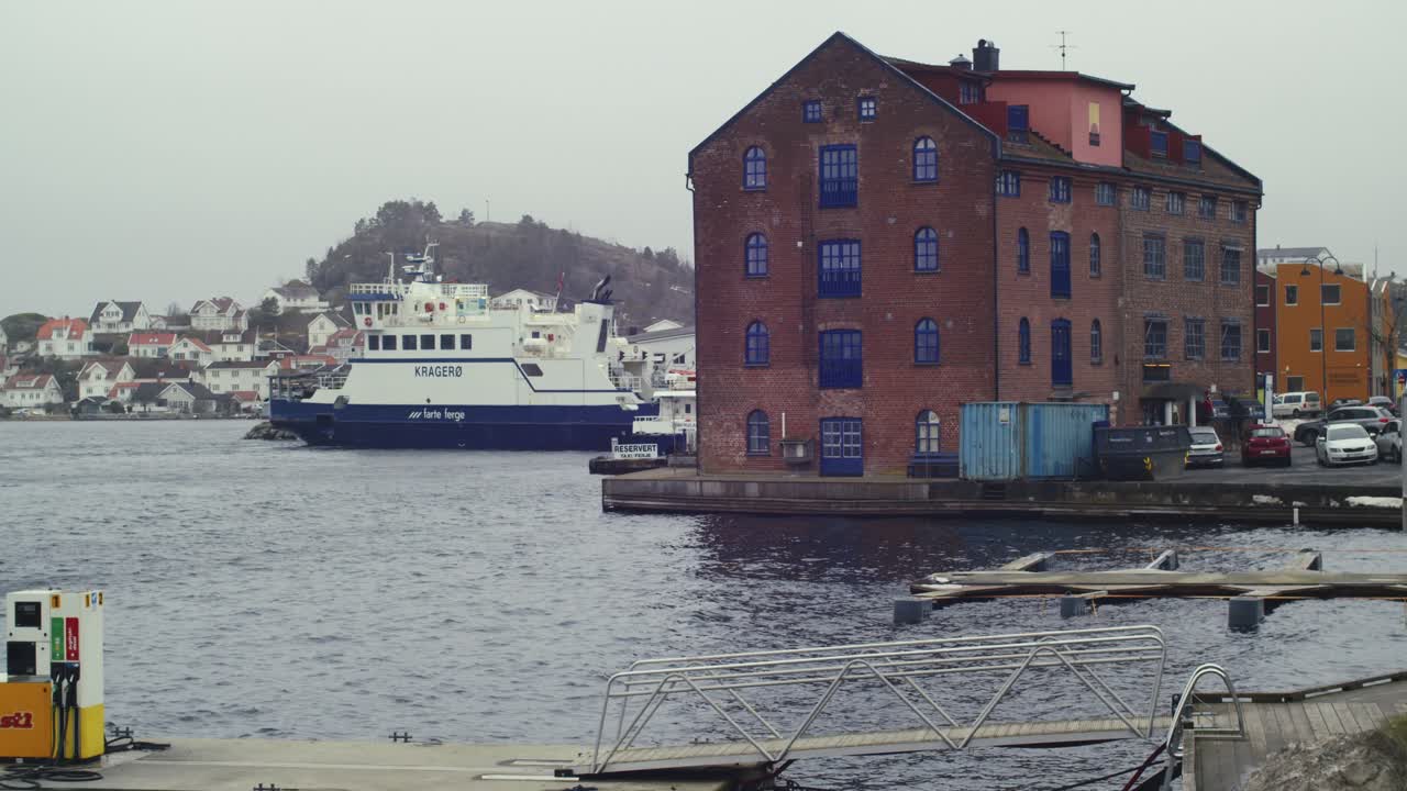 En statisk panoramavy av en jättelik lastfärja och en stor tegelbyggnad i Kragerø, Telemark, Norge, vid en bensinstation, en molnig dag.