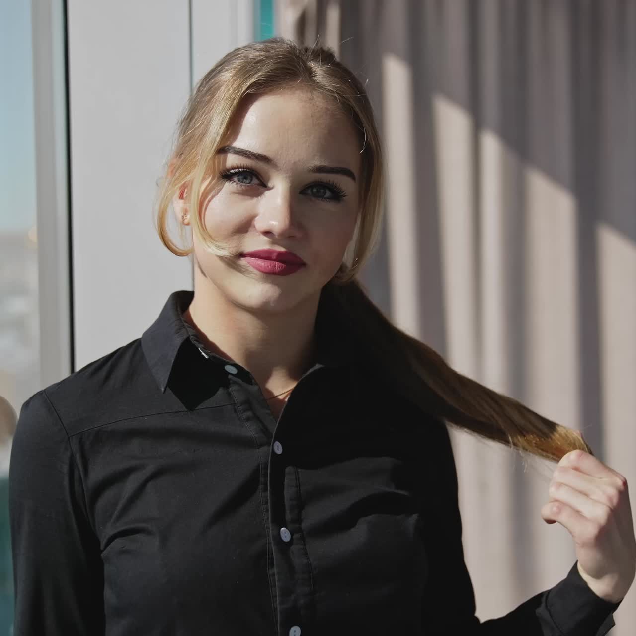 Beautiful young employee in a sunlit office posing in front of the camera. Business lady wearing dark shirt looks straight into the camera. Woman moving closer to the camera