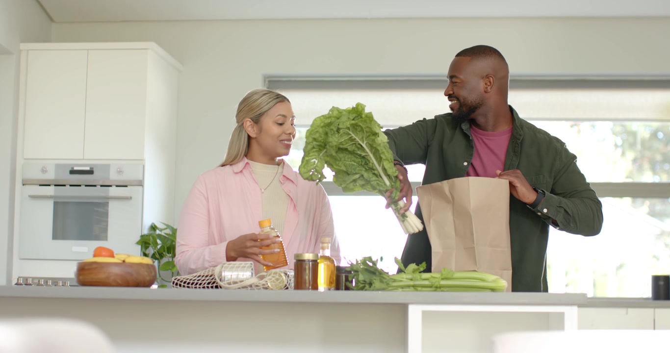 Couple déballant une livraison de courses sur l'îlot de cuisine, sortant des légumes verts d'un sac en papier pour préparer les repas.