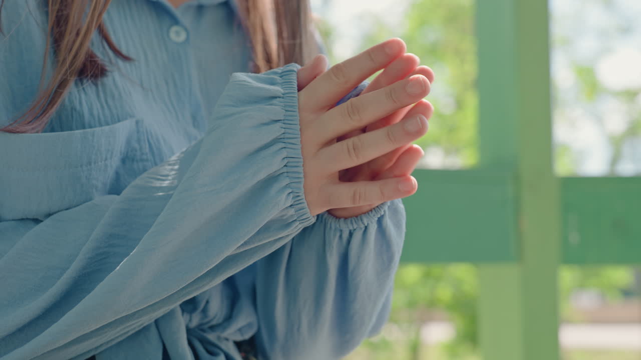 녹색 벤치에 앉아 두 손을 비비고 감싸 쥐는 백인 여성, 긴 파란 소매가 살짝 당겨져 있고, 얼룩덜룩한 햇빛 아래 부드럽게 온기를 느끼는 몸짓, 공원 배경과 섬세한 손의 디테일이 돋보여요