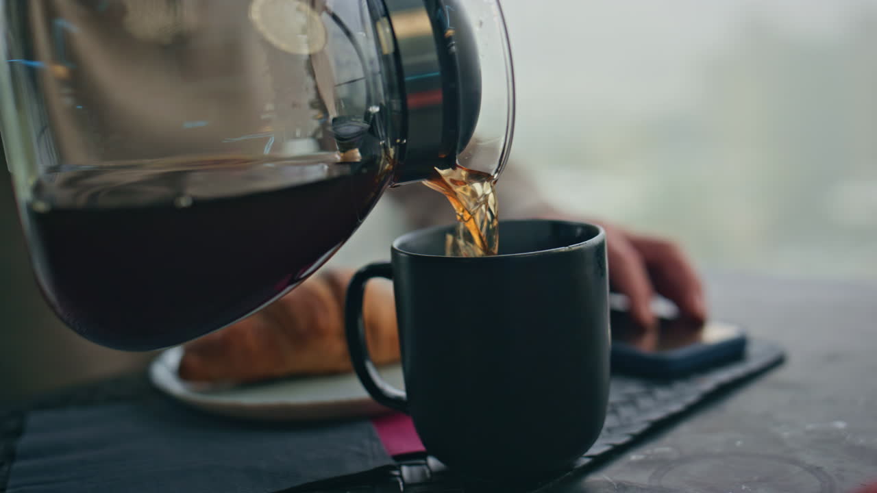 человек с руками, наливающим чашку кофе, большие окна, кухня вблизи. человек, смотрящий телефон.