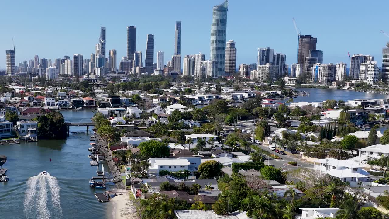 Drone follows motorboat along canal with city skyline, residential homes, bright daylight, smooth motion