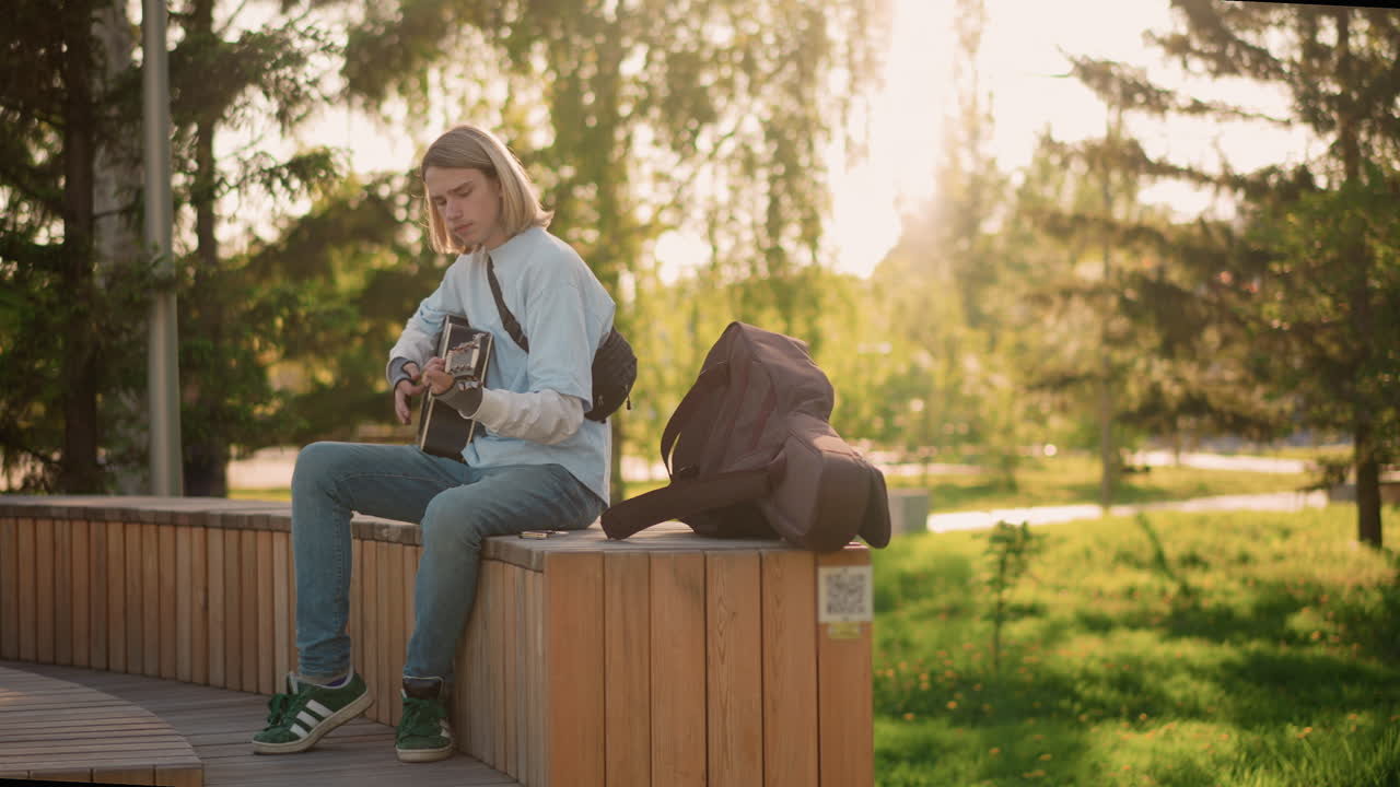 Chitarrista nel parco durante l'ora d'oro che suona musica allegra, strimpellando una chitarra acustica vicino alla custodia aperta, calda retroilluminazione, jeans comodi, passanti attenti, riverberi solari, sfondo naturale, atmosfera estiva vivace