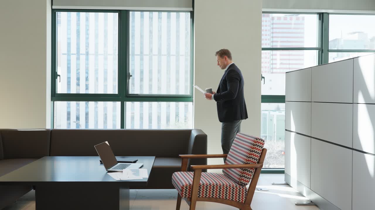 A businessman in a dark suit reviews documents and drinks coffee while standing by large windows in a modern office space featuring a minimalist desk, laptop, and a retro-styled armchair