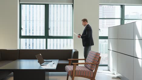 a businessman in a dark suit reviews documents and drinks coffee while standing by large windows in a modern office space featuring a minimalist desk, laptop, and a retro-styled armchair