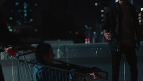 young-group-of-friends-racing-shopping-cart-having-fun-on-rooftop-at-night-enjoying-weekend-celebration-party