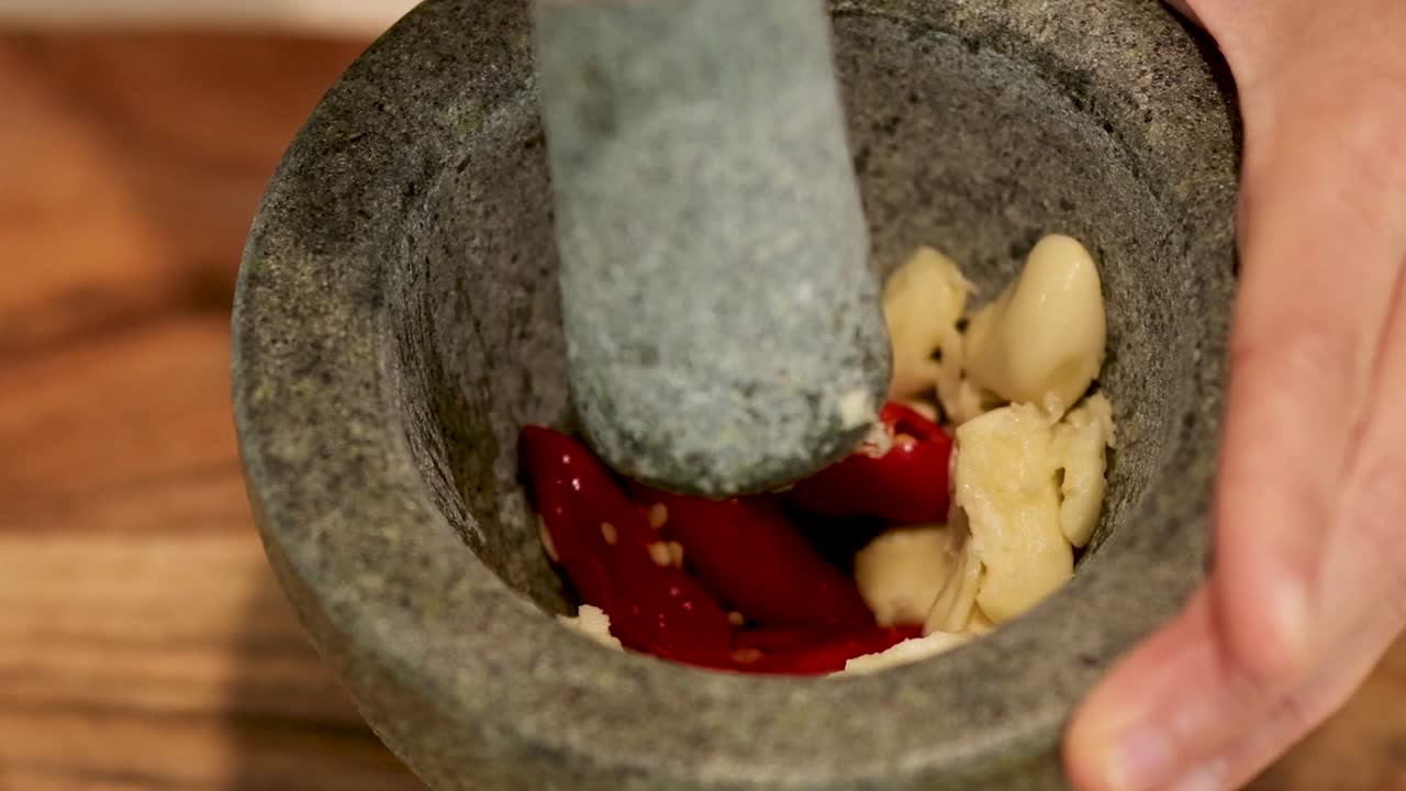 Una persona usa un pestello di pietra per schiacciare manualmente spicchi d'aglio fresco e peperoncini rossi