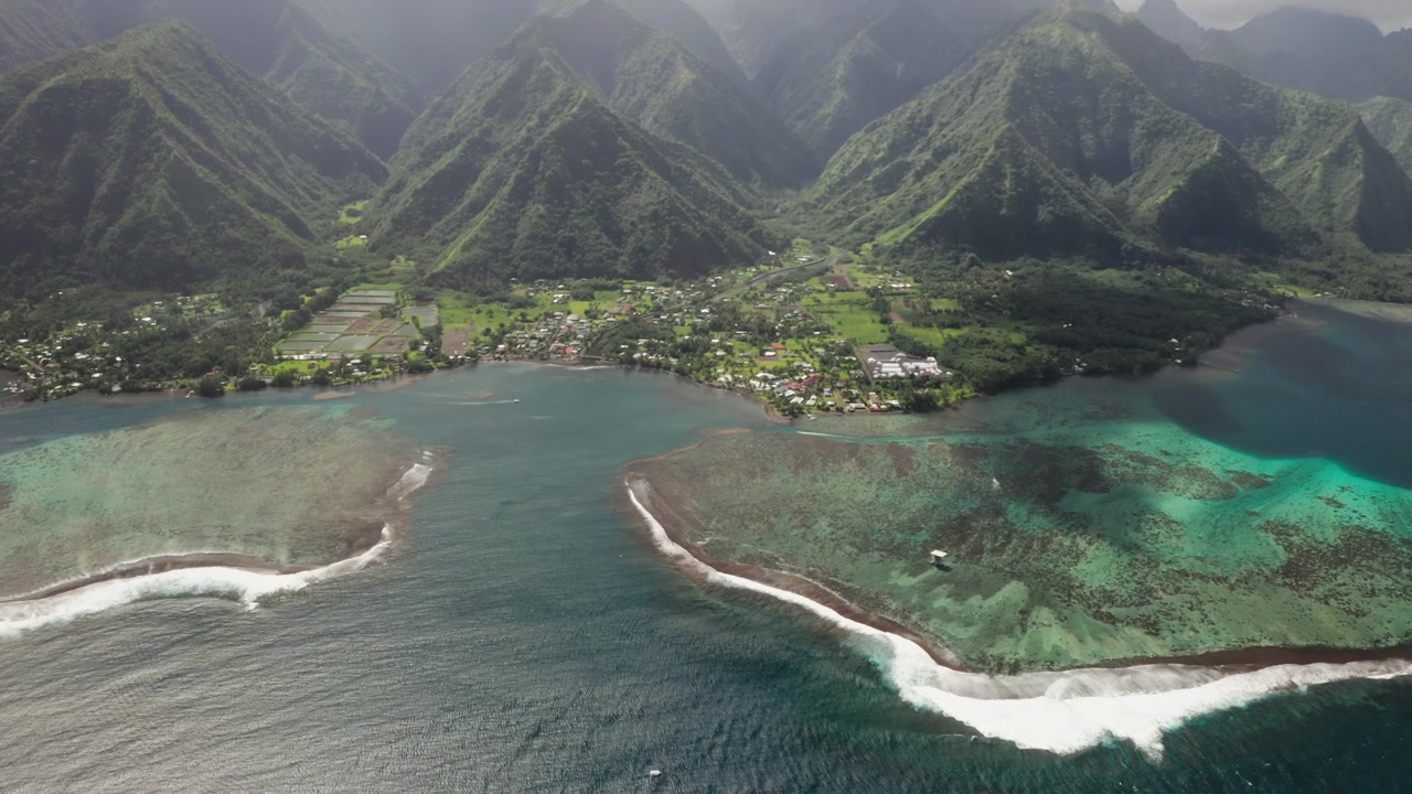 Steady drone shot over Teahupoo village river mouth, Tahiti, calm lagoon water reflecting overcast sky beside scattered rooftops and palm trees.