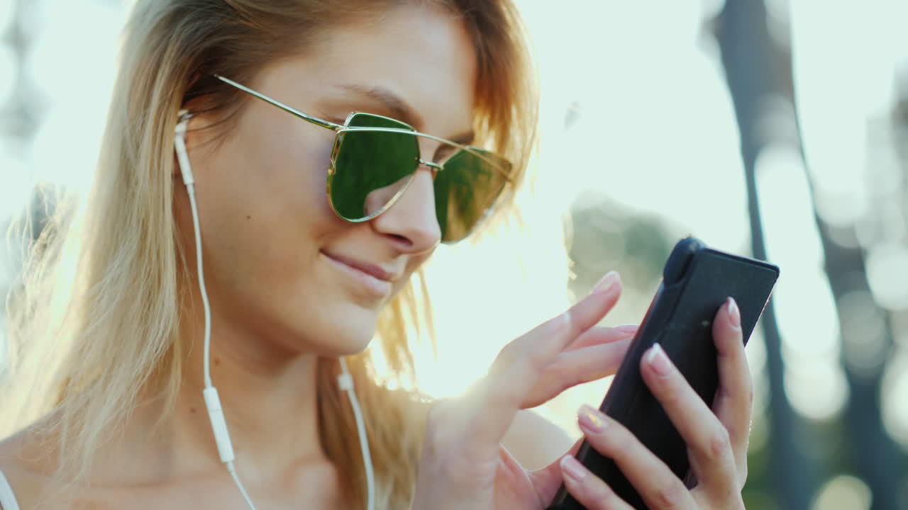 선글라스를 쓴 세련된 여성은 휴가 중에 스마트폰을 사용하여 태양 광선을 아름답게 비춥니다