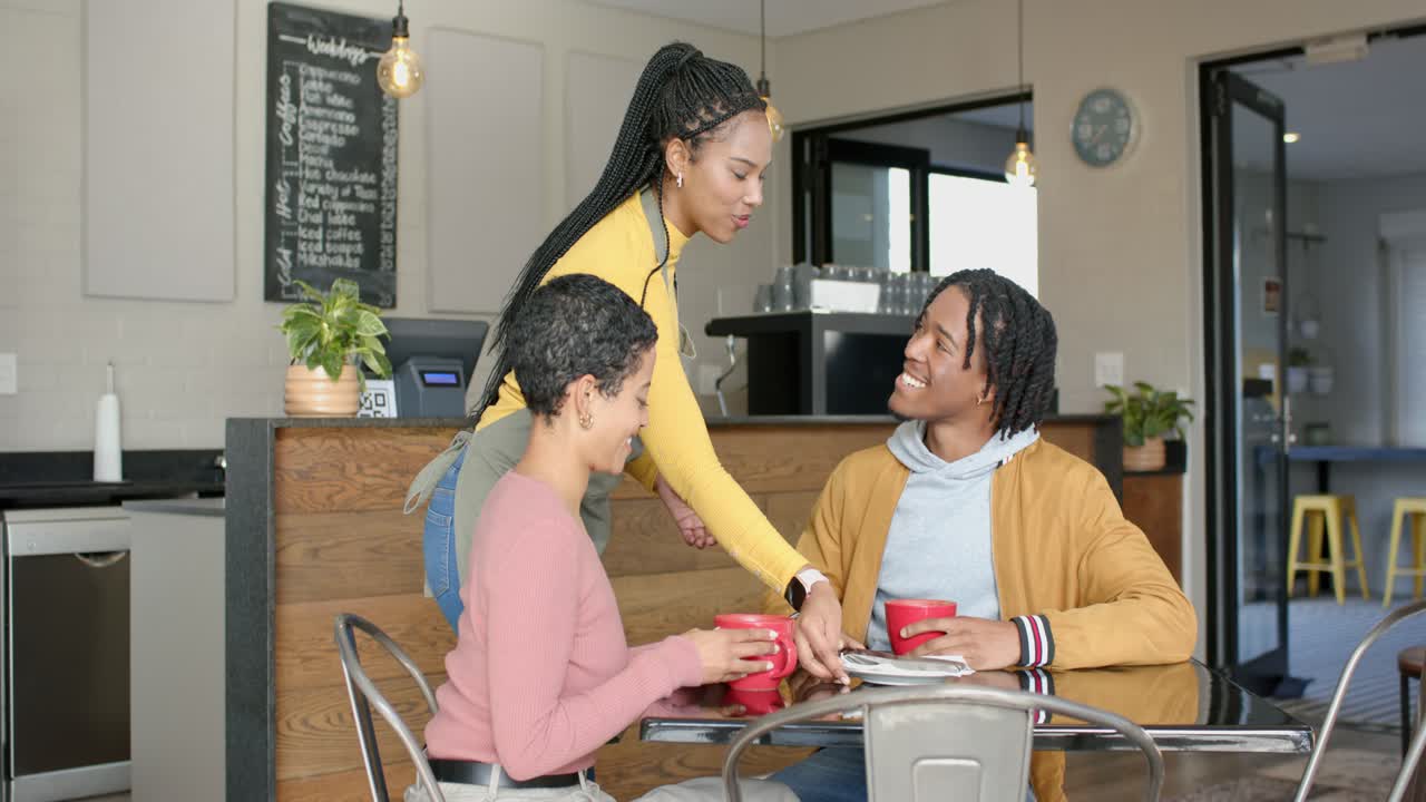 Female waitress carrying tray serving plate, African American couple smiling with red mugs at cafe