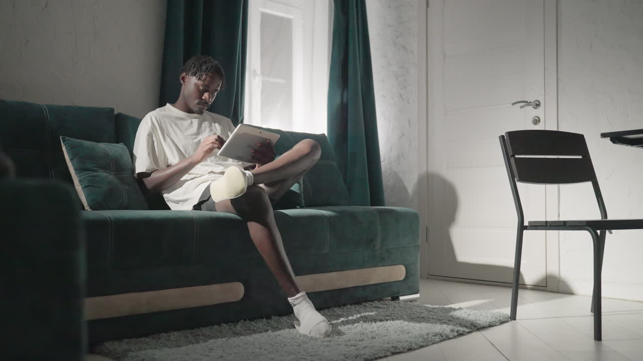 Champ seated on teal sofa, reading tablet with full attention. Natural light filters in through window, creating a calm and peaceful atmosphere in modern, cozy living room setting