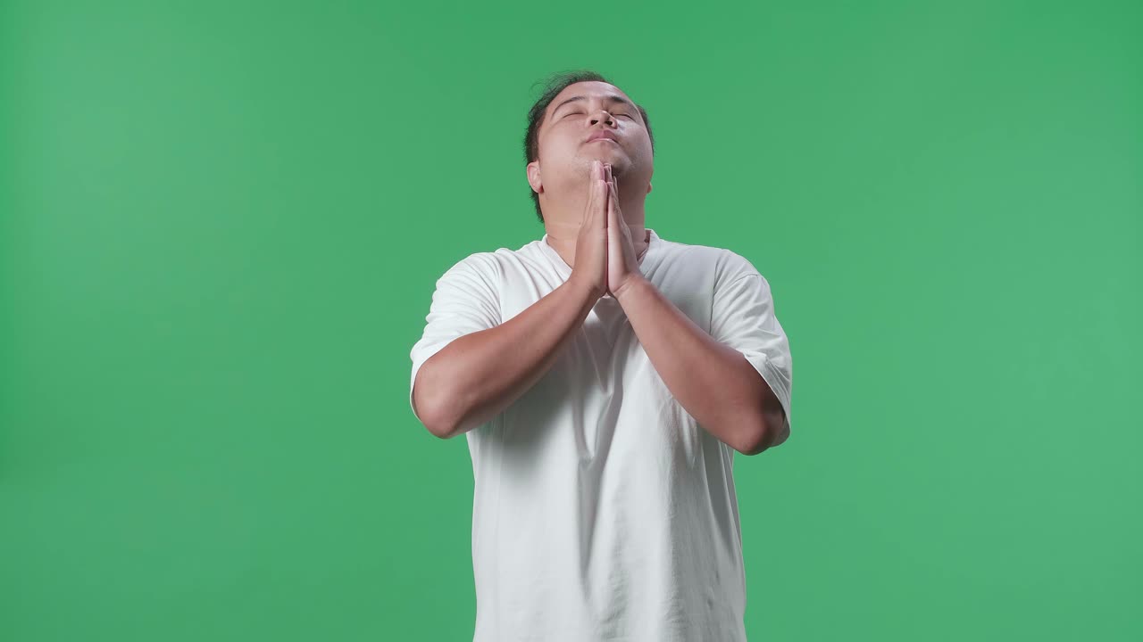 눈을 감고 공을 들고 있는 아시아인 남자가 초록색 스크린 배경에서 축구를 응원하는 동안 손을 들기 전에 팀을 위해 기도합니다.