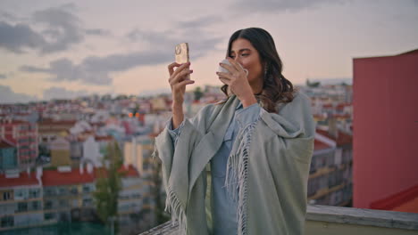 relaxed brunette taking photo sipping tea on balcony with dusk cityscape closeup