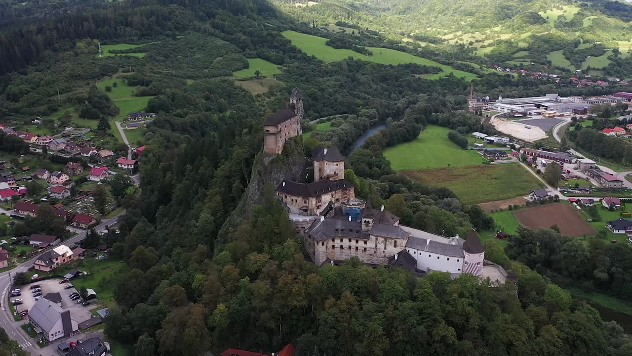 A drone approaches Orava Castle, flying toward its steep rocky foundations and revealing the castle’s striking silhouette against the Slovak countryside