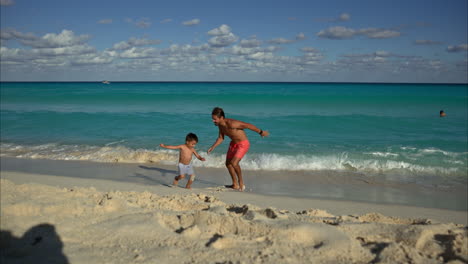 Cámara-Lenta-De-Un-Hombre-Latino-Mexicano-Jugando-Con-Su-Hijo-En-La-Playa-Huyendo-De-Las-Olas-En-Una-Playa-De-Cancún