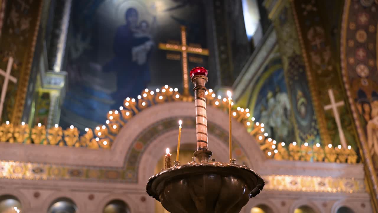 A low-angle view of the sacred art inside St. Volodymyr's Cathedral in Kyiv, Ukraine. Warm candlelight glows on ornate church ornaments, with a historic fresco and cross in the background.