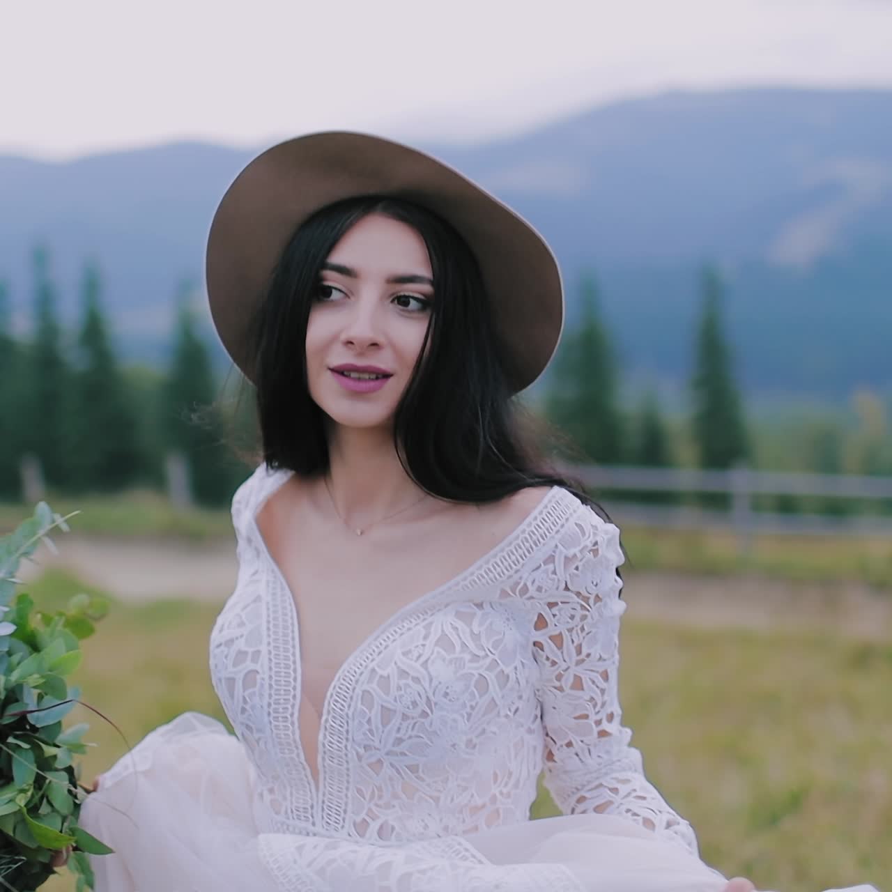 Portrait of lovely dark-haired bride in nature. Beautiful woman in stylish wedding dress and a hat posing on camera in the mountainous area.