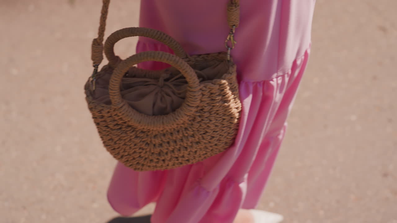 Primo piano di una donna caucasica che cammina su un marciapiede illuminato dal sole con una borsa di paglia, un abito rosa svolazzante e ballerine bianche, mentre una borsa intrecciata oscilla con movimento aggraziato, texture boho e atmosfera estiva calda