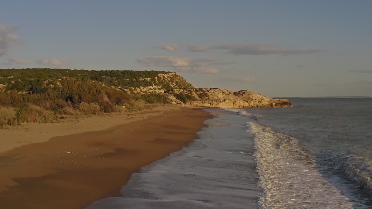 Sorvolo aereo cinematografico di una bellissima spiaggia di sabbia dorata mentre le onde si infrangono sulla riva al tramonto