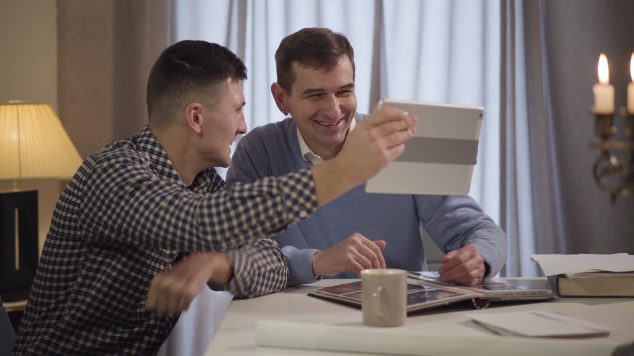 웃는 성인 백인 남자가 중년 아버지에게 비디오 통화를 하는 측면 뷰 초상화. 행복한 아들과 아빠가 저녁에 실내에서 태블릿을 사용합니다. 현대 기술, 라이프 스타일, 세대