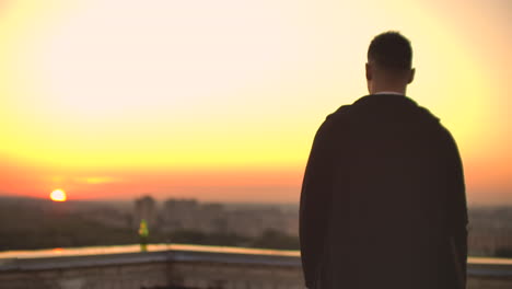 back view a male freelance programmer in a hoodie walks on the roof at sunset and drinks beer looking at the beautiful view of the city.