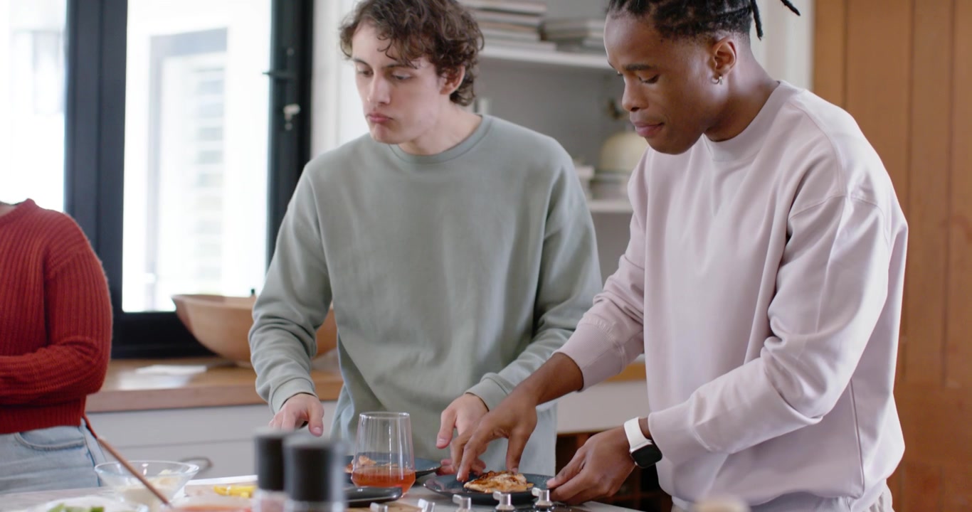 Two male friends tasting then plating at kitchen island, preparing meal, arranging utensils