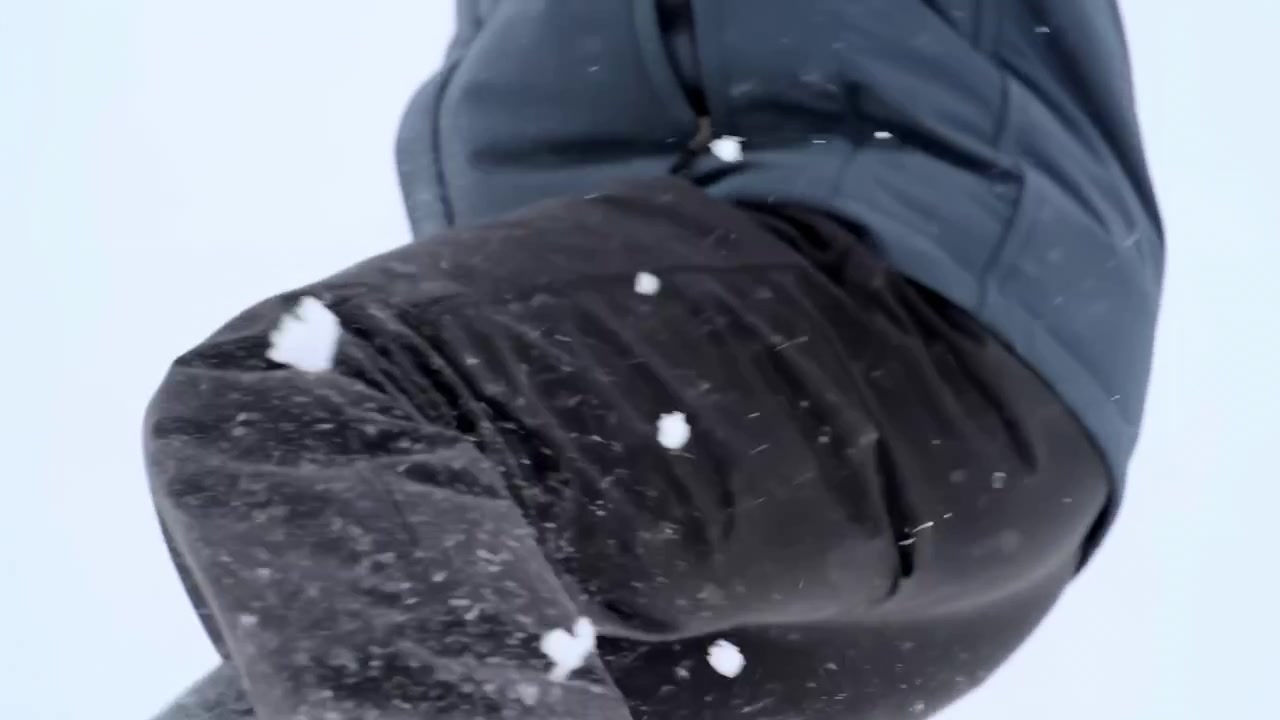 Tight cropped view of lower torso and bent knee in dark insulated outerwear while airborne snow spray and white clumps strike fabric, creating visible winter impact, texture, and rugged outdoor energy