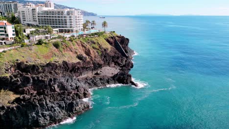 Tiro-Hundido-Desde-La-Línea-De-La-Costa-Con-Hoteles-Y-Horizonte-En-Tiro-Hacia-El-Mar-Que-Muestra-Las-Olas-ásperas-Rompiendo-Contra-Las-Rocas-Costeras