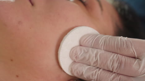 close up of spa visitor face with gloved hand using soft wool pad to rub cheek to forehead during soothing facial cleansing treatment in bright spa clinic setting with soft ambient lighting
