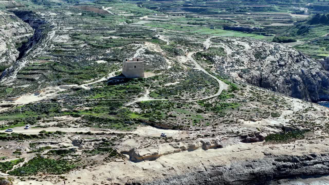 Parlak güneşli bir bahar gününde, canlı yeşillik ve toprak tonlu kıyı manzarası eşliğinde Gozo Malta'nın engebeli kireçtaşı kayalıklarını koruyan Tarihi Dwejra Gözetleme Kulesi'nin havadan yüksek açılı görünümü.