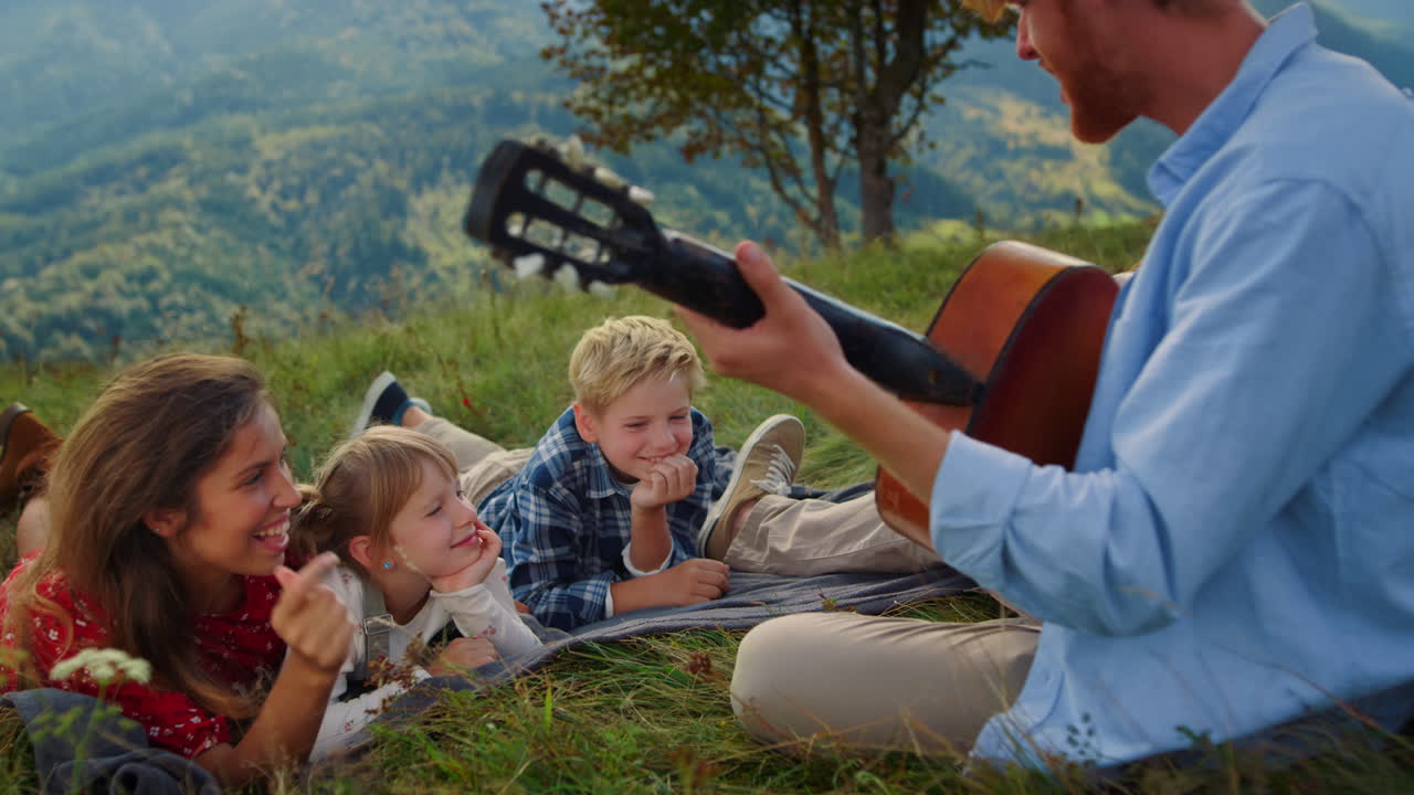 Familie lytter til guitarmusik på grøn bakke. Far spiller for kvinde med børn.