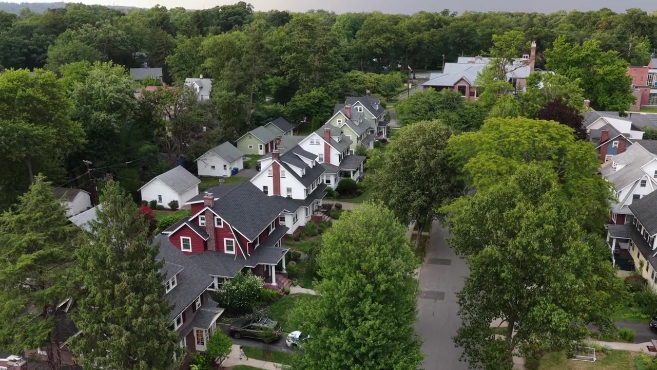 Case storiche fiancheggiano una strada residenziale alberata e frondosa a South Orange, New Jersey, mostrando tetti a capanna, camini in mattoni e portici avvolgenti sotto un cielo tempestoso. Ampia ripresa aerea con drone in ascesa