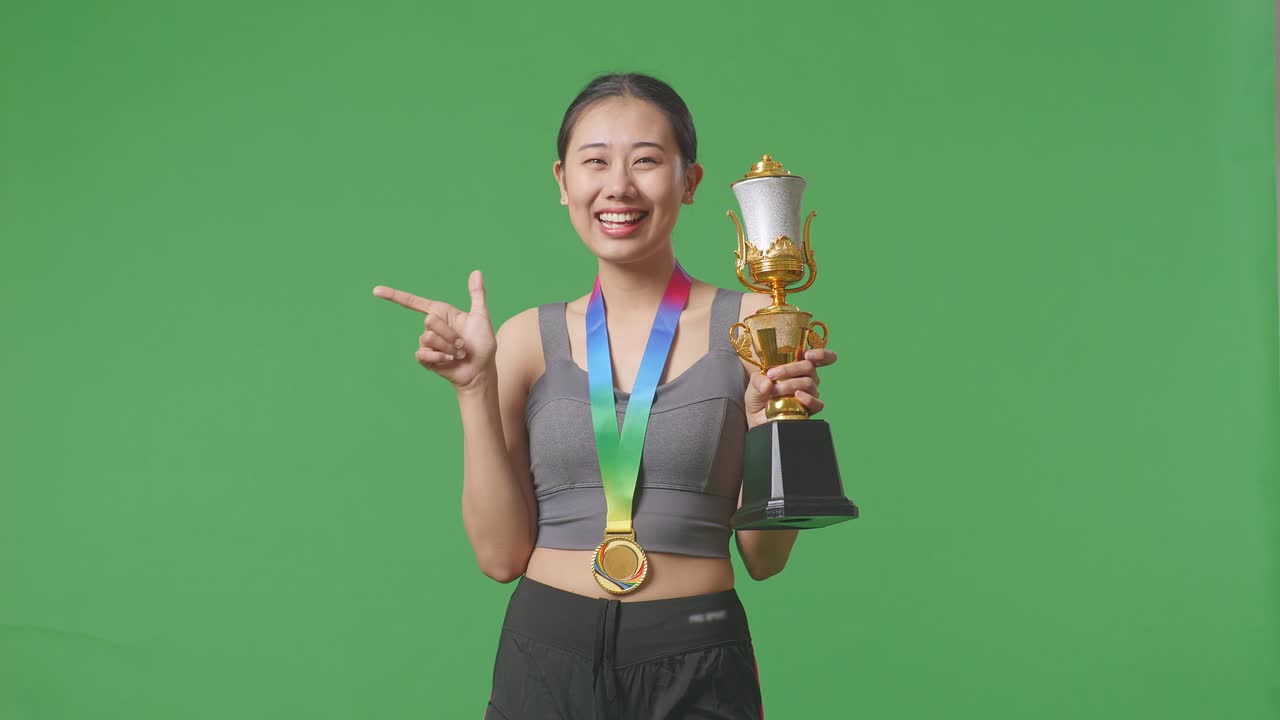 금메달을  아시아 여성이 손에 금 트로피를 들고 미소 짓고 스튜디오의 녹색 스크린 배경에 을 가리키고 있습니다.