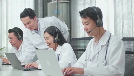 a man coming to help a woman of three asian doctors in headsets using computer for working as call center agent while her colleagues are speaking and typing during a call at the office