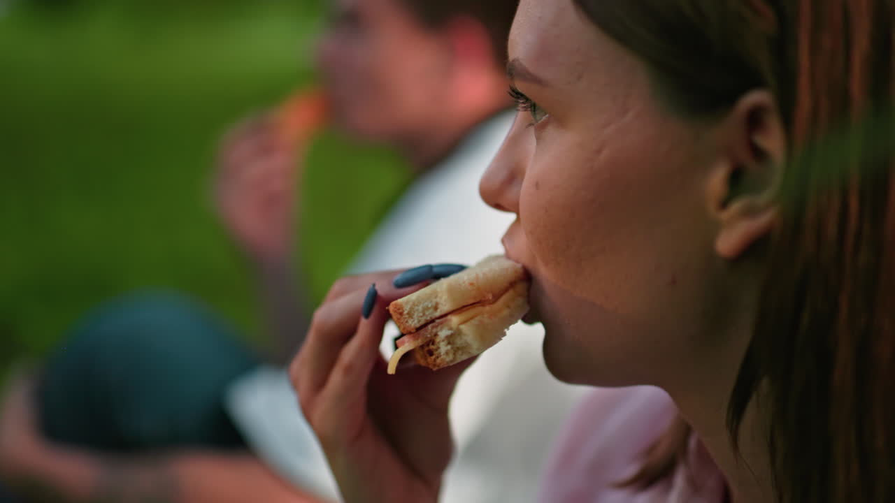 primer plano de una mujer con uñas pulidas comiendo un sándwich a la parrilla en la mano, con un fondo borroso que muestra a alguien con una camiseta blanca masticando, un entorno al aire libre relajado con iluminación suave