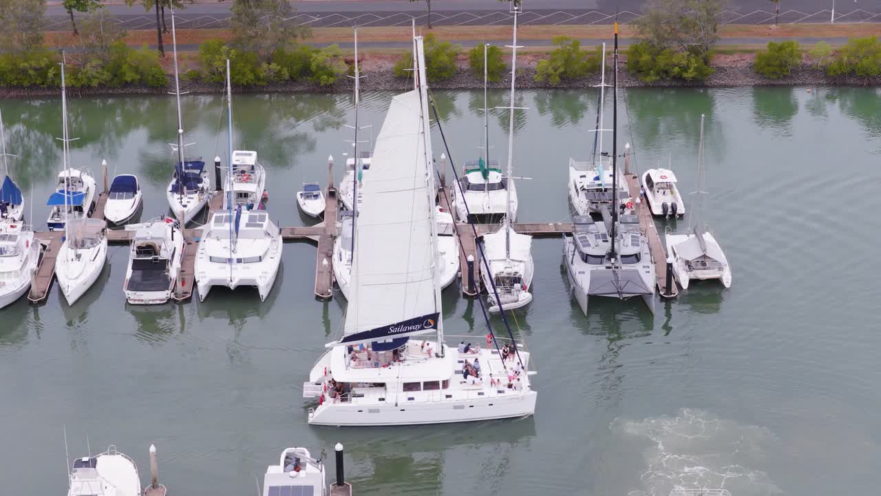A sailboat maneuvers through a marina, surrounded by docked boats under soft daylight in Port Douglas, Australia