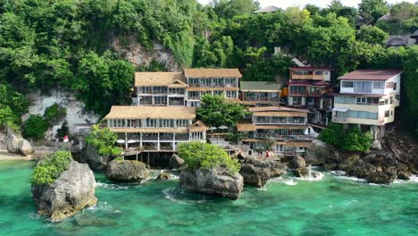 turquoise ocean waves crashing on large rocky coastline with homes on a cliff, aerial