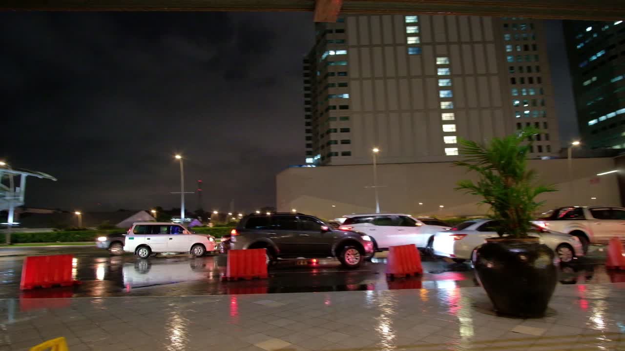 Outdoor evening scene showing vehicular traffic in a drizzle around a commercial district within Quezon City in Metro Manila, Philippines.