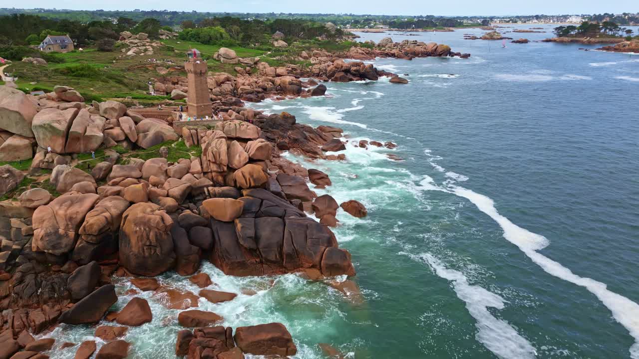 Reverse drone shot revealing the Mean Ruz lighthouse on the Pink Granite Coast in Ploumanac'h. Waves crash on the reddish rocks with greenery and coastline in background - Brittany France