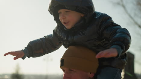 a close-up of a father carrying his son on his shoulders playfully as the boy stretches his arms out, the father wears a brown jacket, and the son wears a black jacket