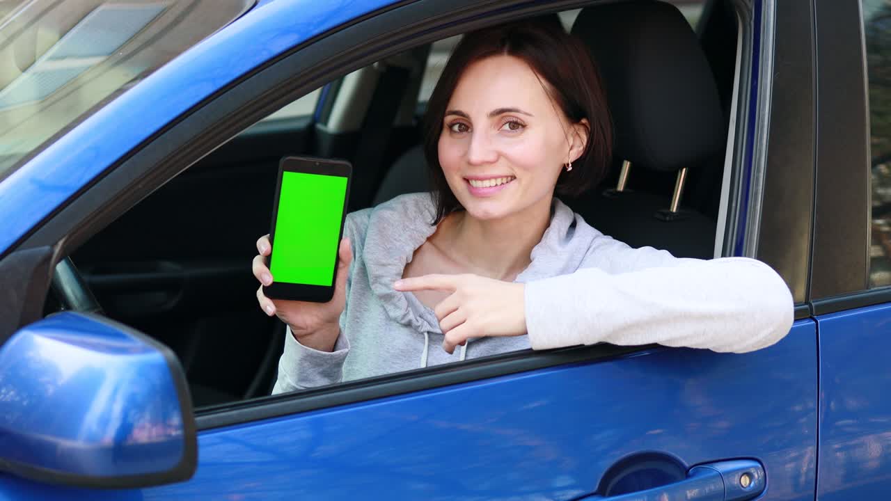 mujer europea con cabello corto en ropa gris sentada en el coche azul, sonriendo y mostrando un teléfono móvil con pantalla verde